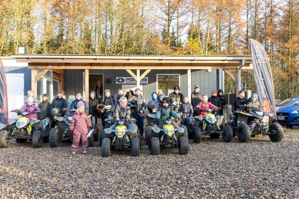 Stor gruppe børn og unge med deres ATV-maskiner foran klubhuset på en solrig dag